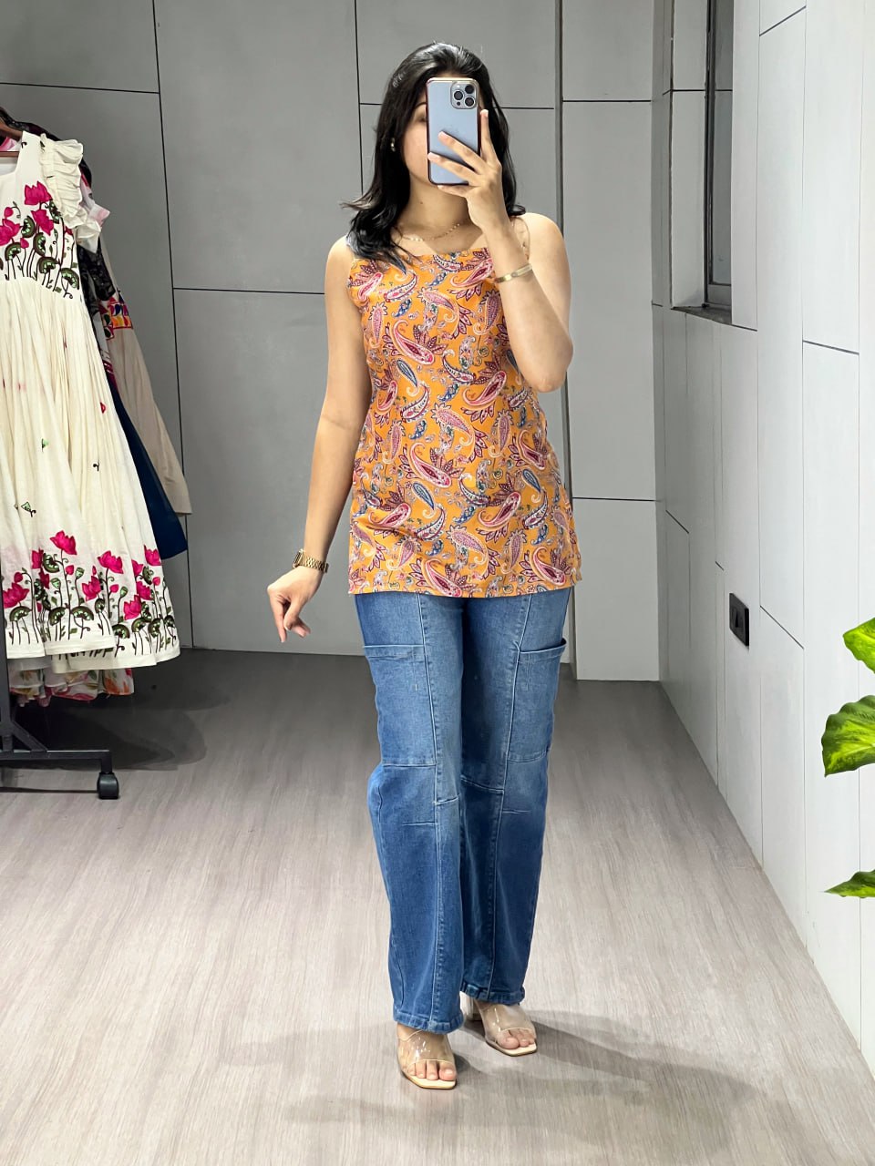 Person wearing a patterned sleeveless top and blue jeans in a room with a wardrobe and plant.