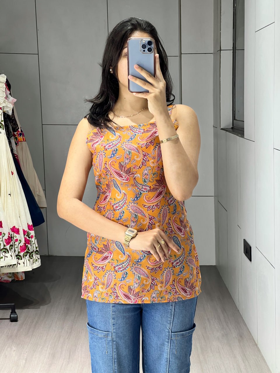 Person taking a mirror selfie wearing a patterned top and blue jeans in a room with gray walls and a white wardrobe.