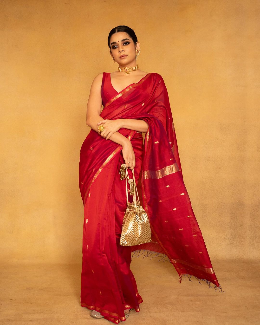 Woman in a red saree with gold accents holding a gold bag against