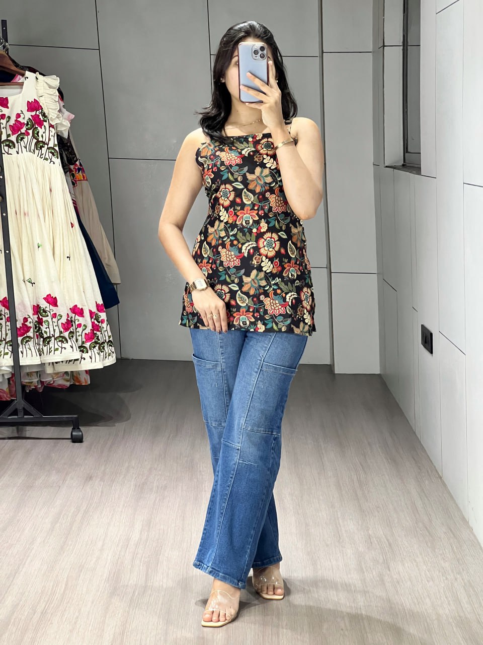 Woman taking a mirror selfie wearing a floral top and blue jeans in a room.