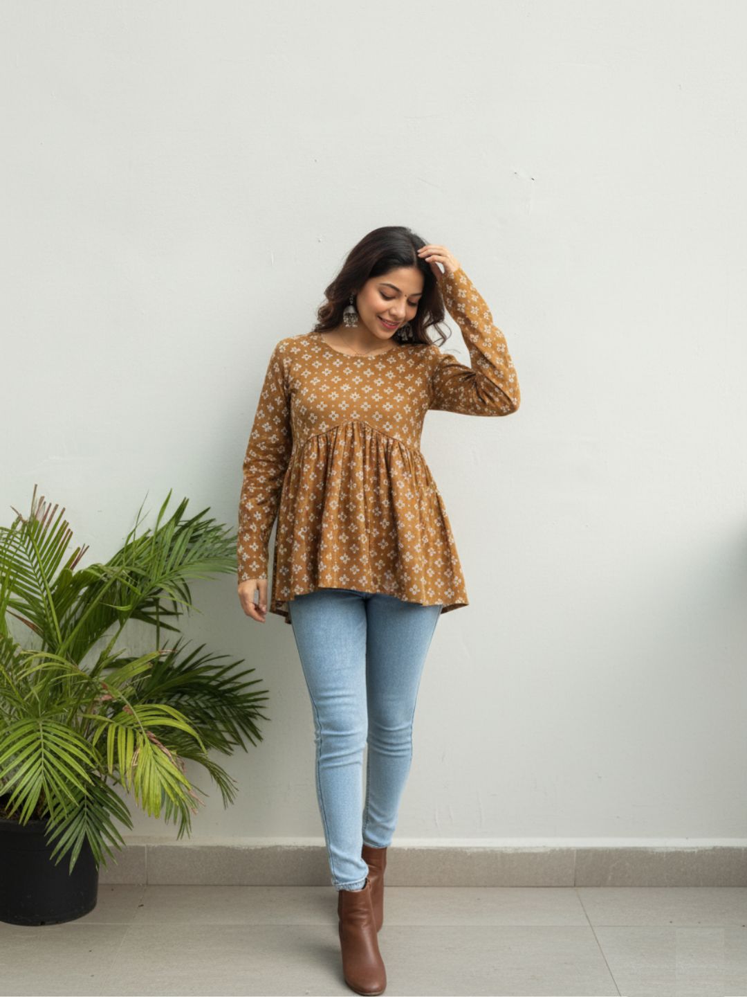 Woman wearing a patterned brown top and light blue jeans standing indoors with a plant in the background.