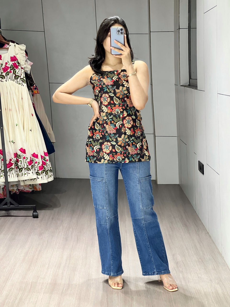 Person wearing a floral top and blue jeans taking a mirror selfie in a fitting room.