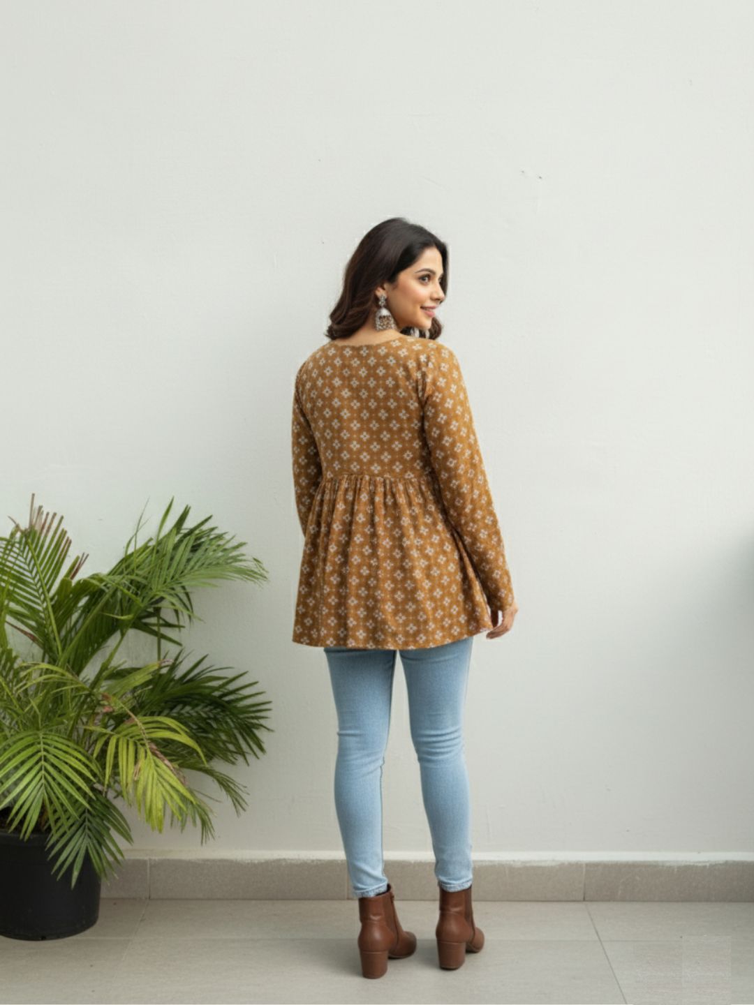 Woman wearing a patterned brown top and light blue jeans standing against a white wall with a plant to the left.