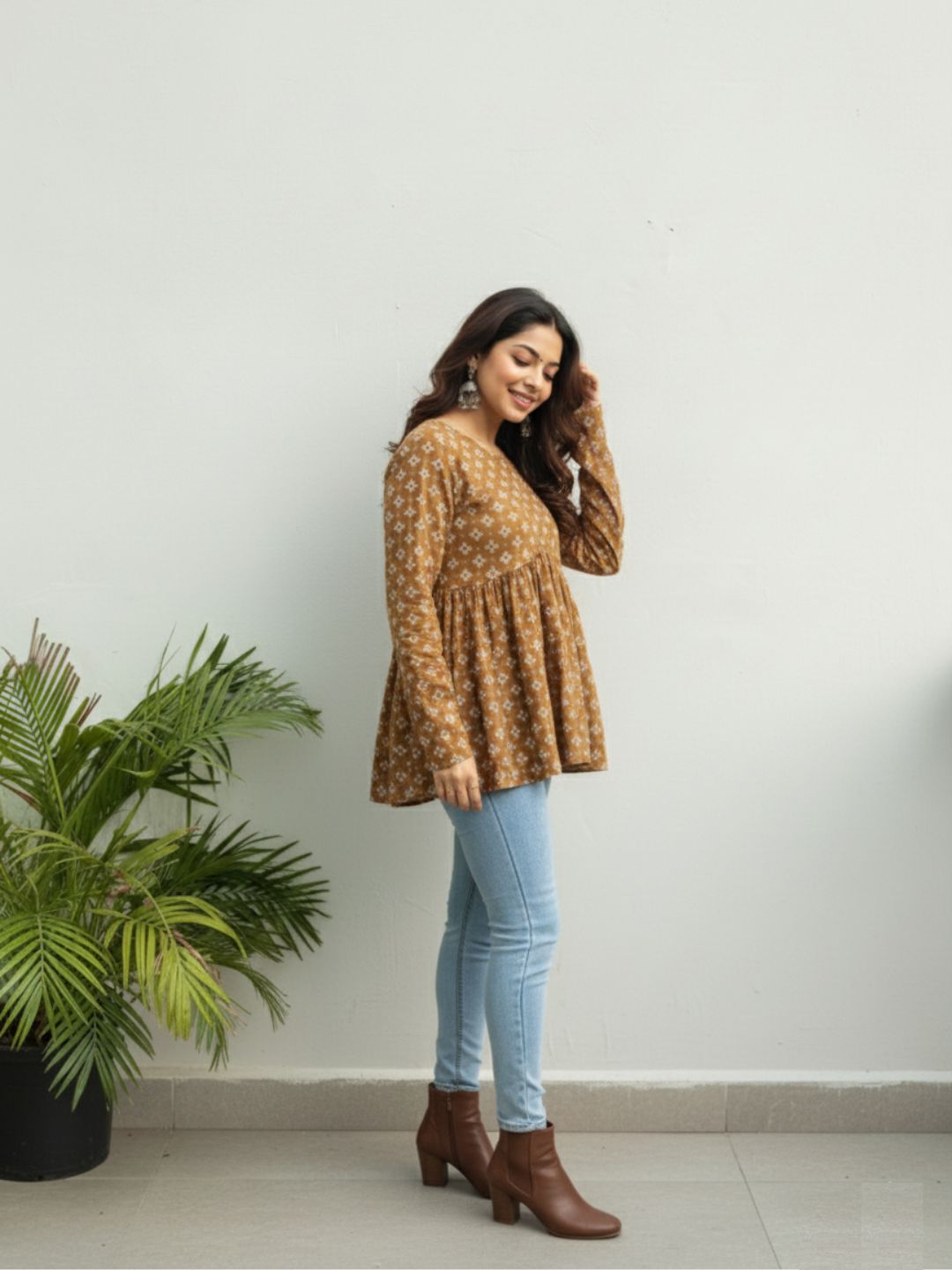 Woman wearing a patterned top and jeans standing next to a plant indoors.