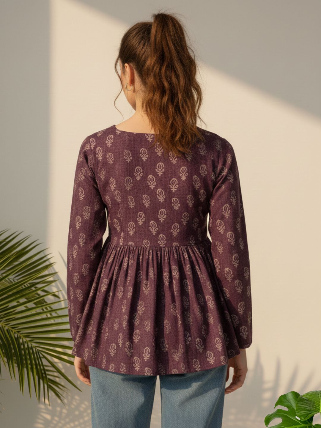 Woman wearing a patterned purple dress standing against a light wall with greenery.