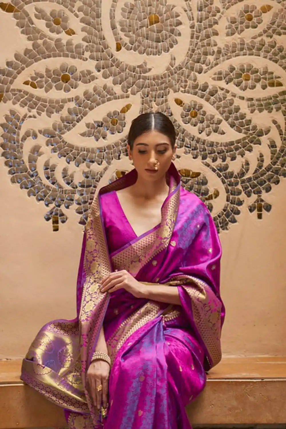 Woman in a pink saree with gold border sitting against a decorative wall.