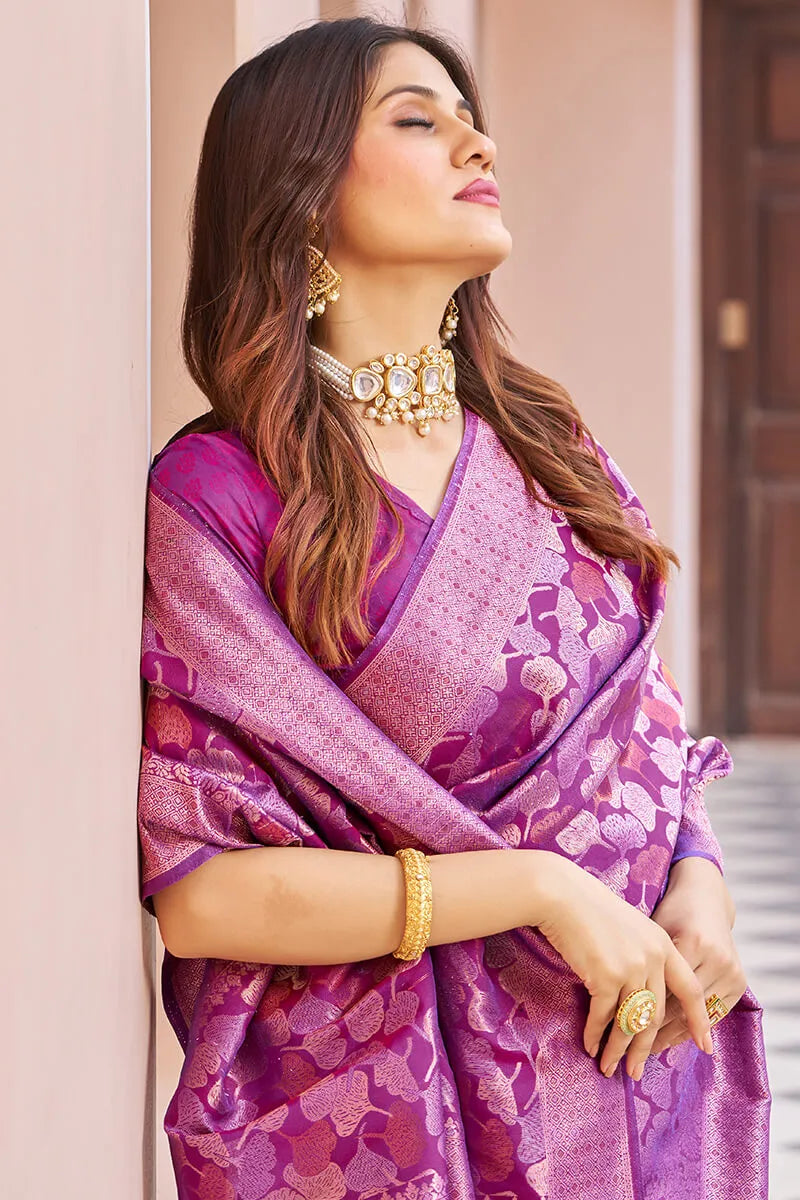 Woman wearing a purple saree with floral patterns, gold jewelry, and a neutral background.