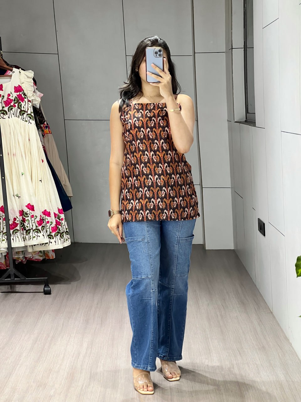 Person wearing a patterned top and blue jeans taking a mirror selfie in a store.