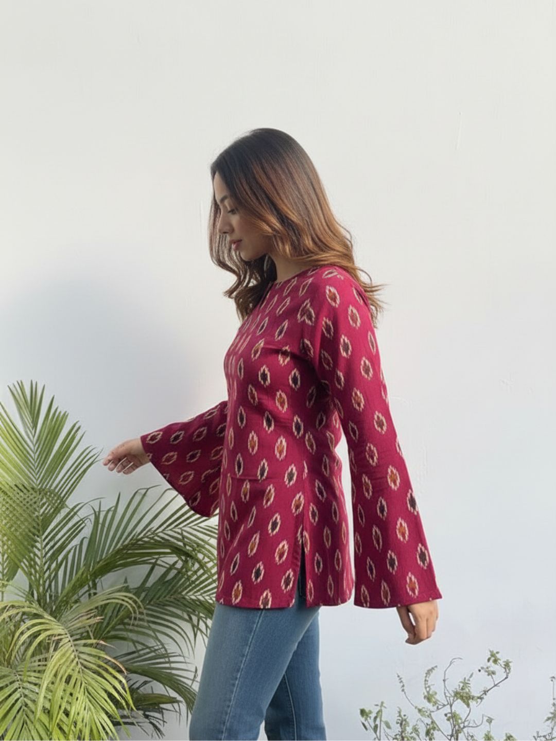 Woman wearing a red patterned blouse with bell sleeves standing next to a plant.