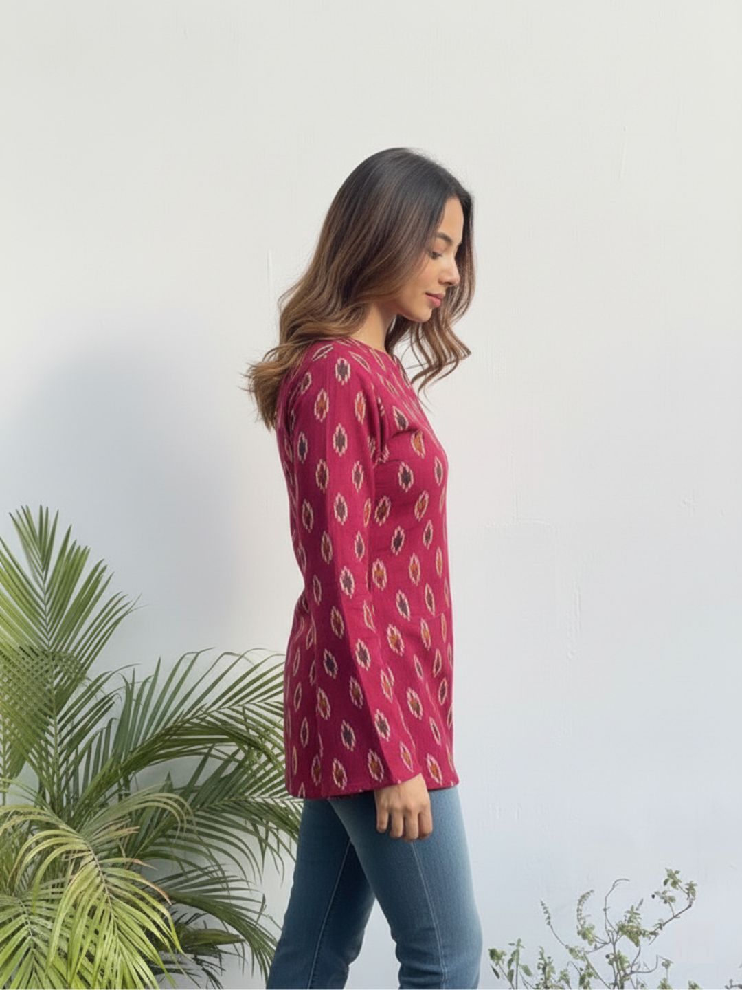 Woman wearing a red patterned top standing next to a plant against a white wall