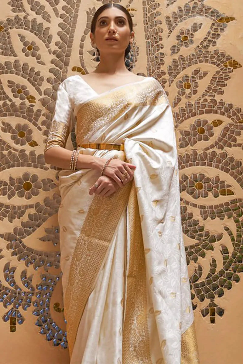 Woman wearing a white and gold saree against a decorative wall.