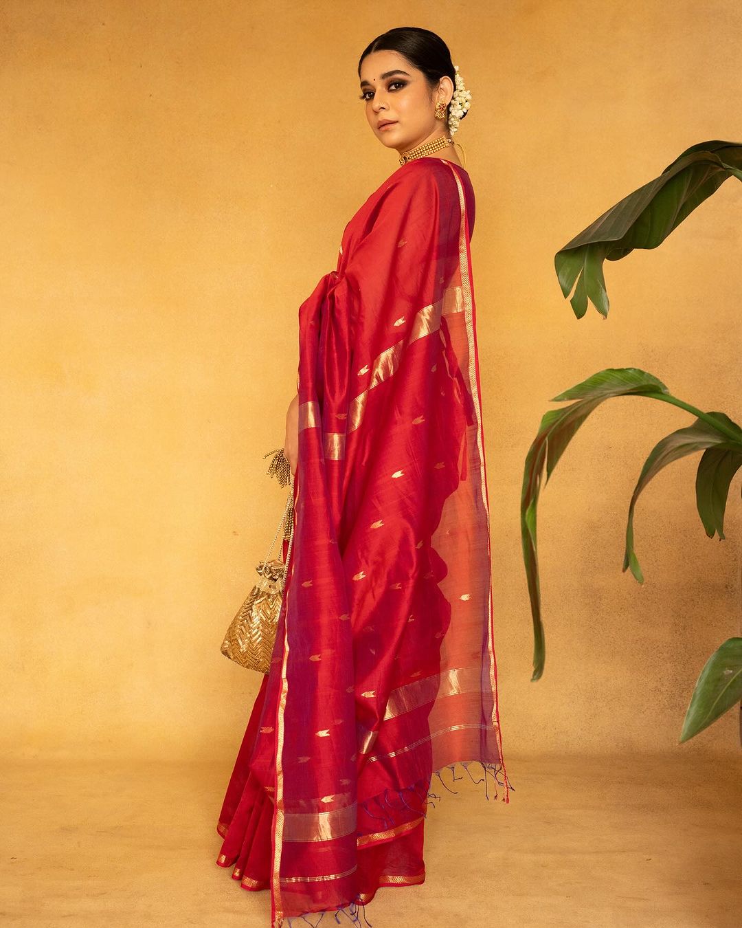 Woman in a red saree with gold accents holding a gold bag against
