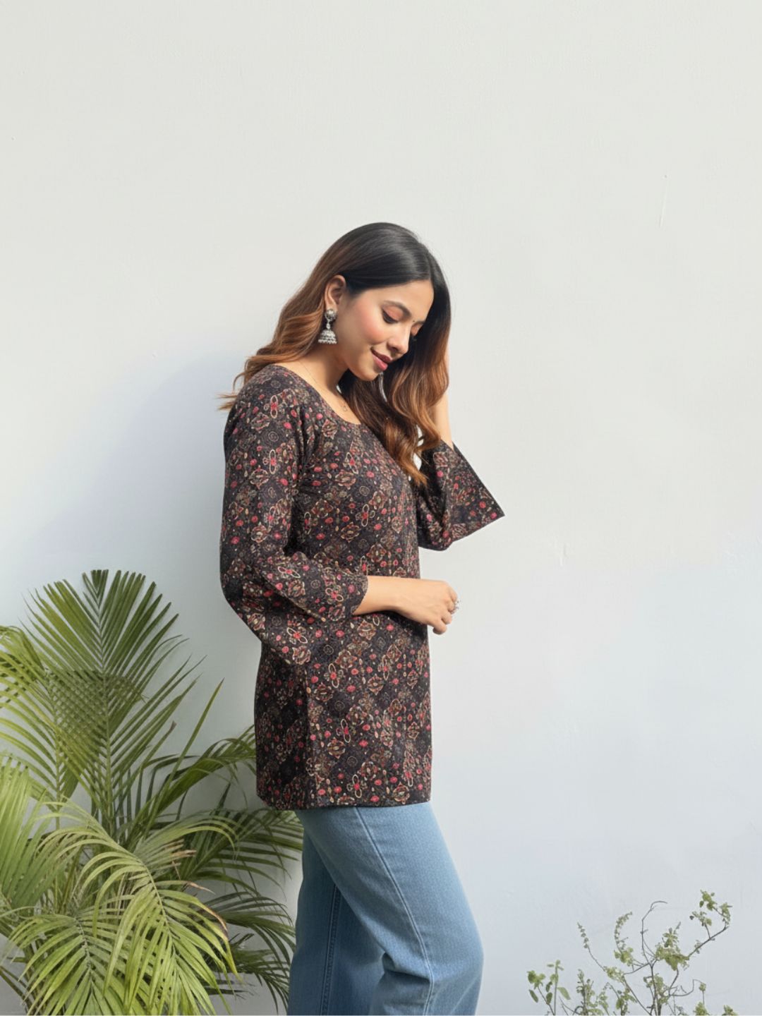 Woman wearing a floral blouse and jeans standing against a light background with plants.