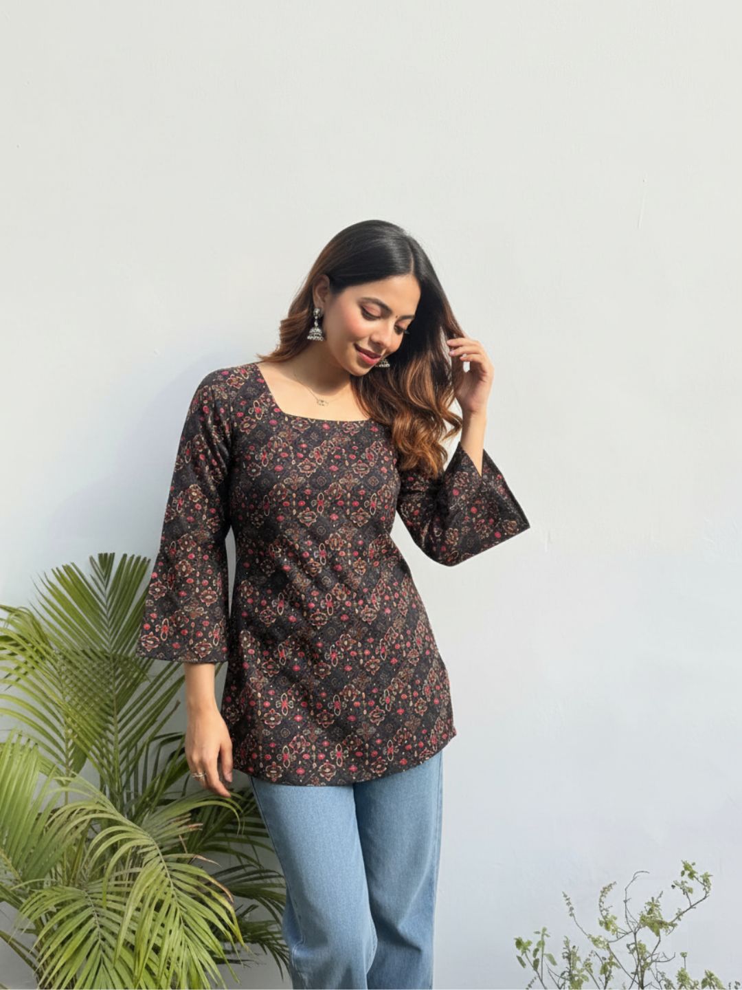 Woman wearing a floral top and blue jeans standing against a light background with plants.