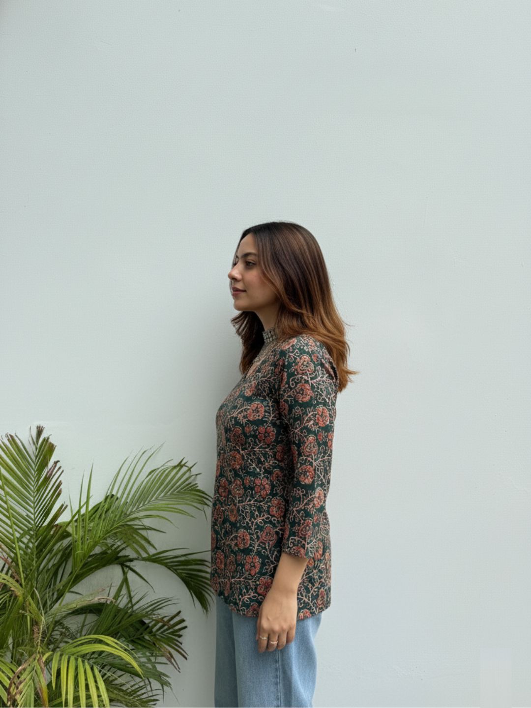 Woman wearing a floral long-sleeve top standing against a light gray wall with a plant to the left.