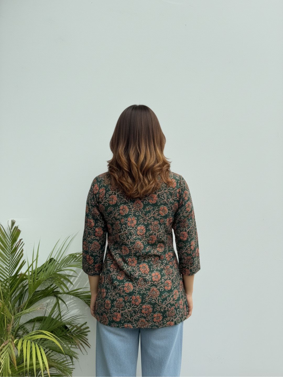 Person wearing a floral patterned jacket standing against a light green wall with a plant to the left.