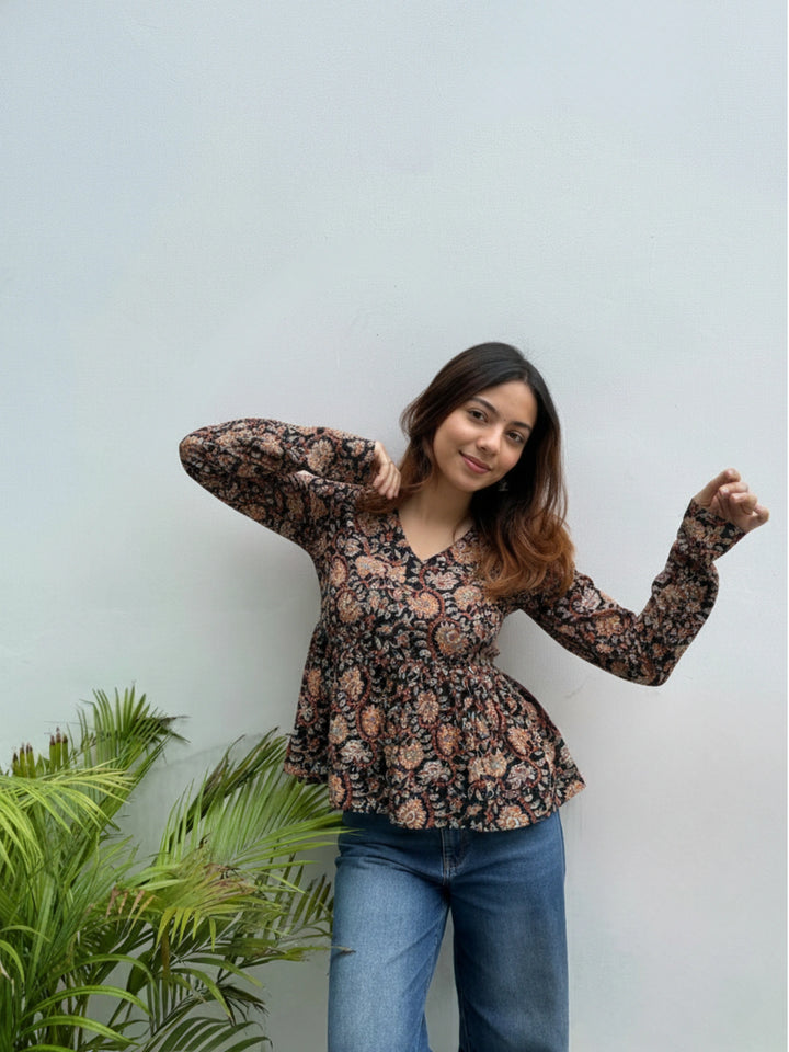 Woman wearing a floral blouse and blue jeans standing against a light green wall with a plant to the left.