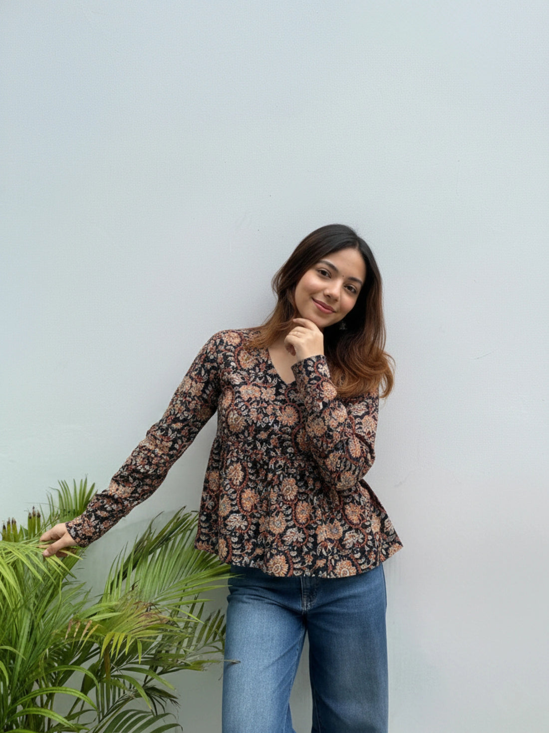 Woman wearing a floral blouse and jeans standing next to a plant against a light blue wall.