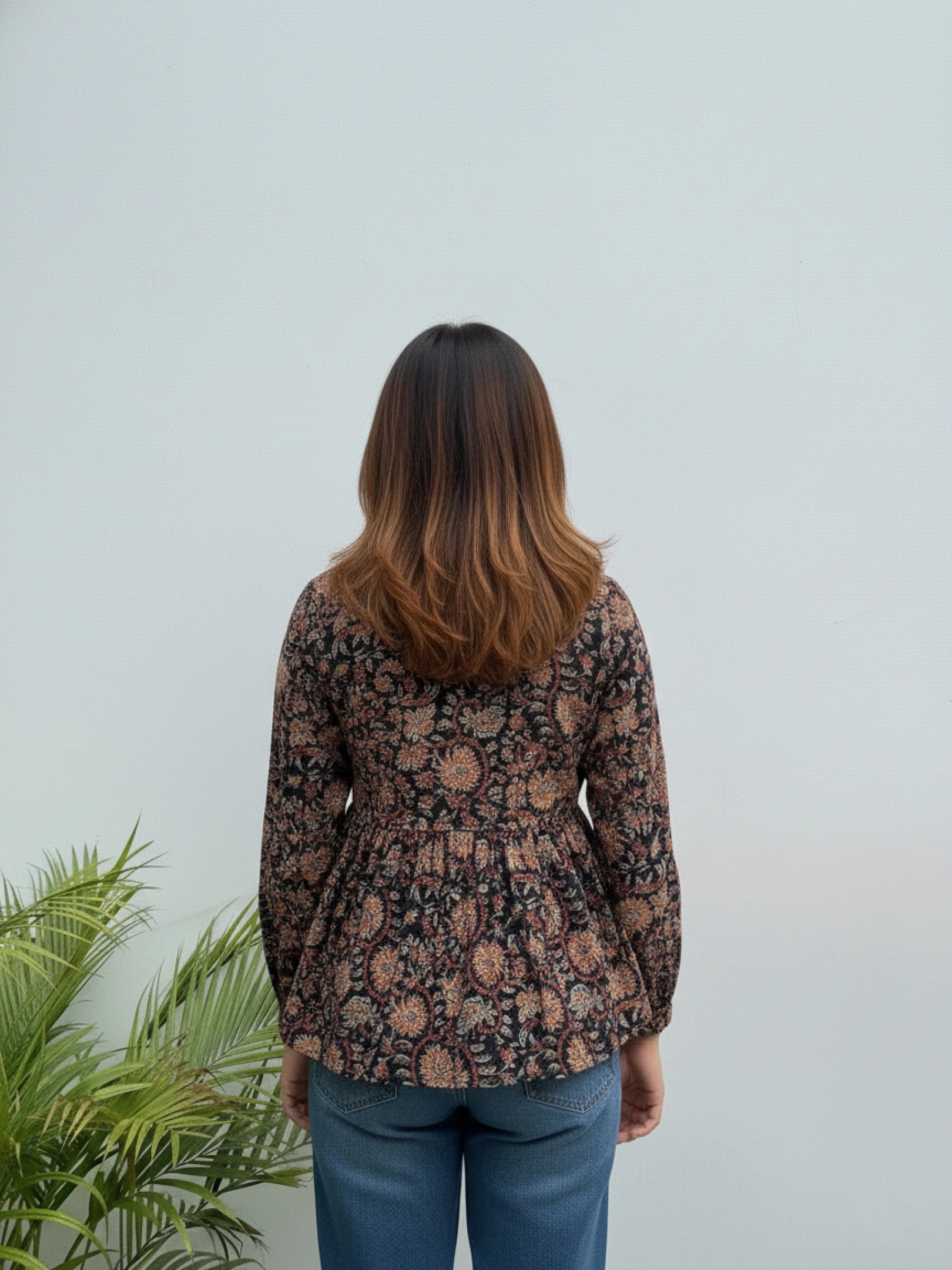 Person wearing a floral blouse standing against a light gray wall with a plant to the left.