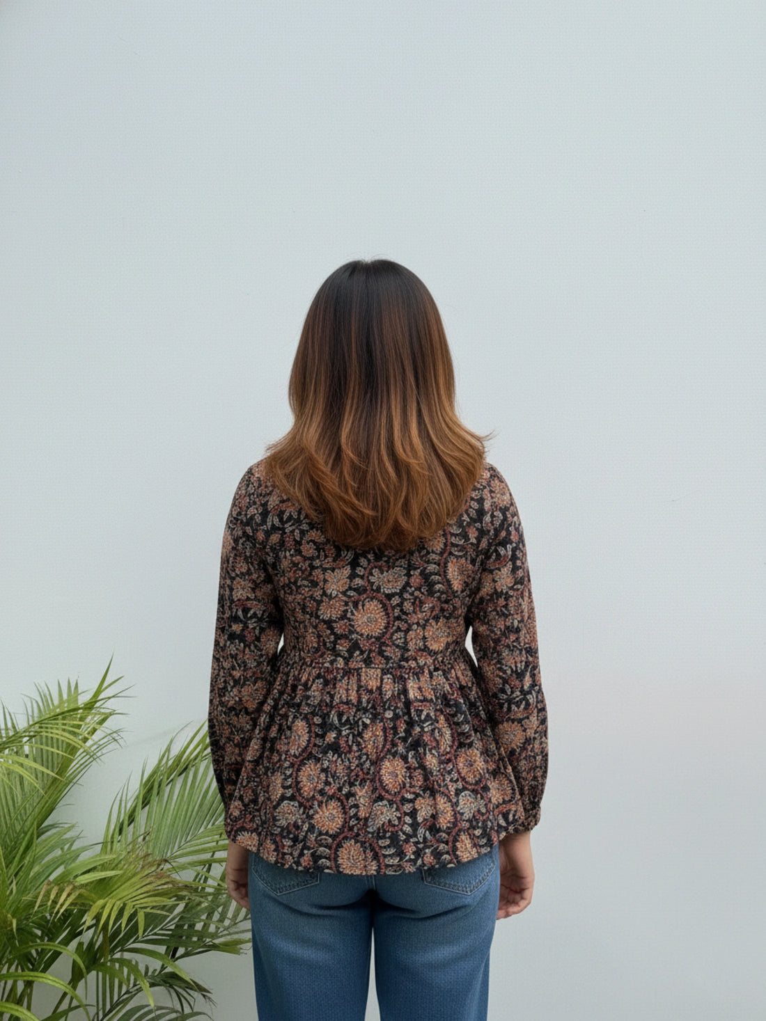 Person wearing a floral blouse standing against a light gray wall with a plant to the left.