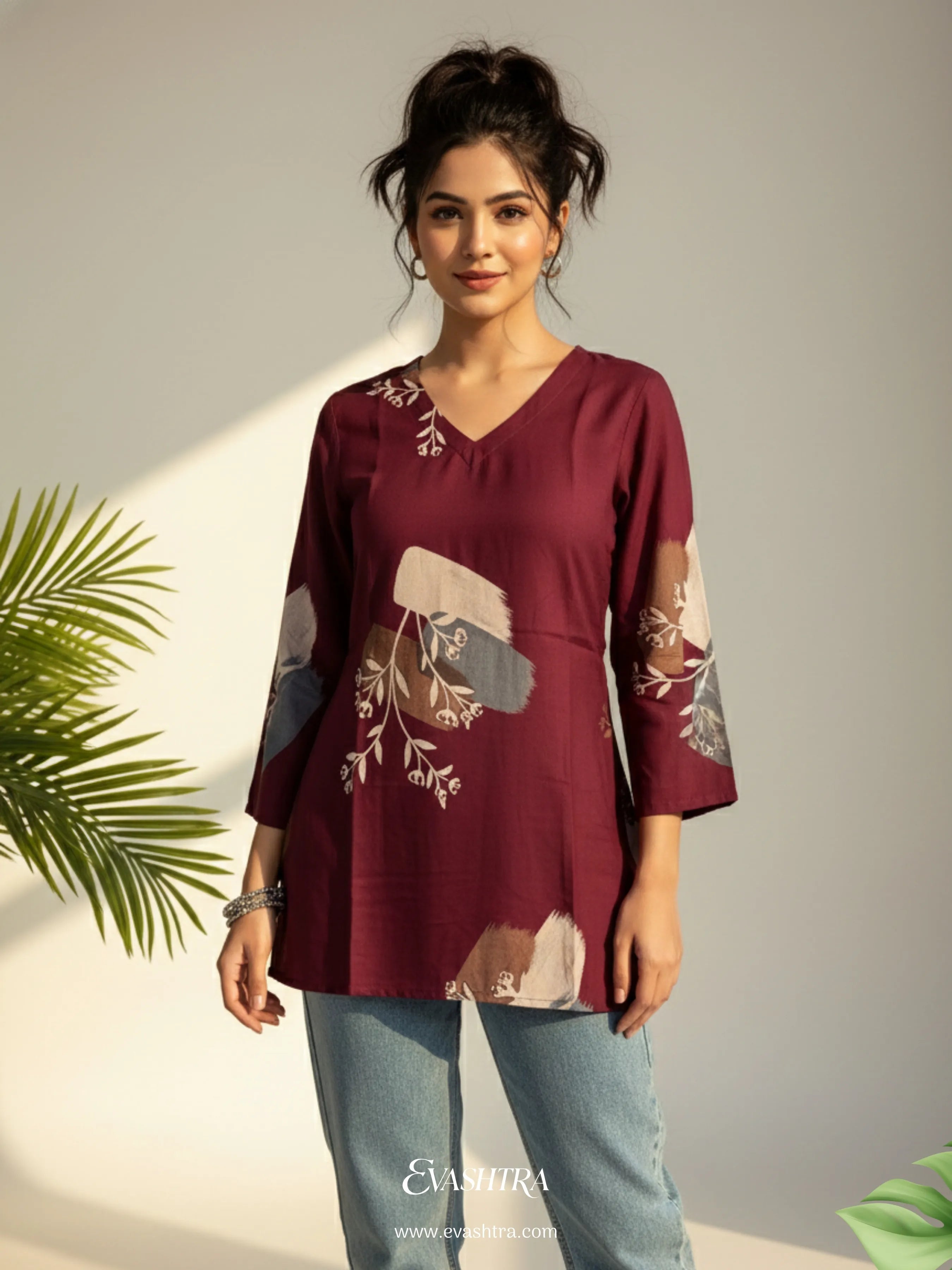 Woman wearing a maroon top with floral patterns and light blue jeans, standing against a neutral background.