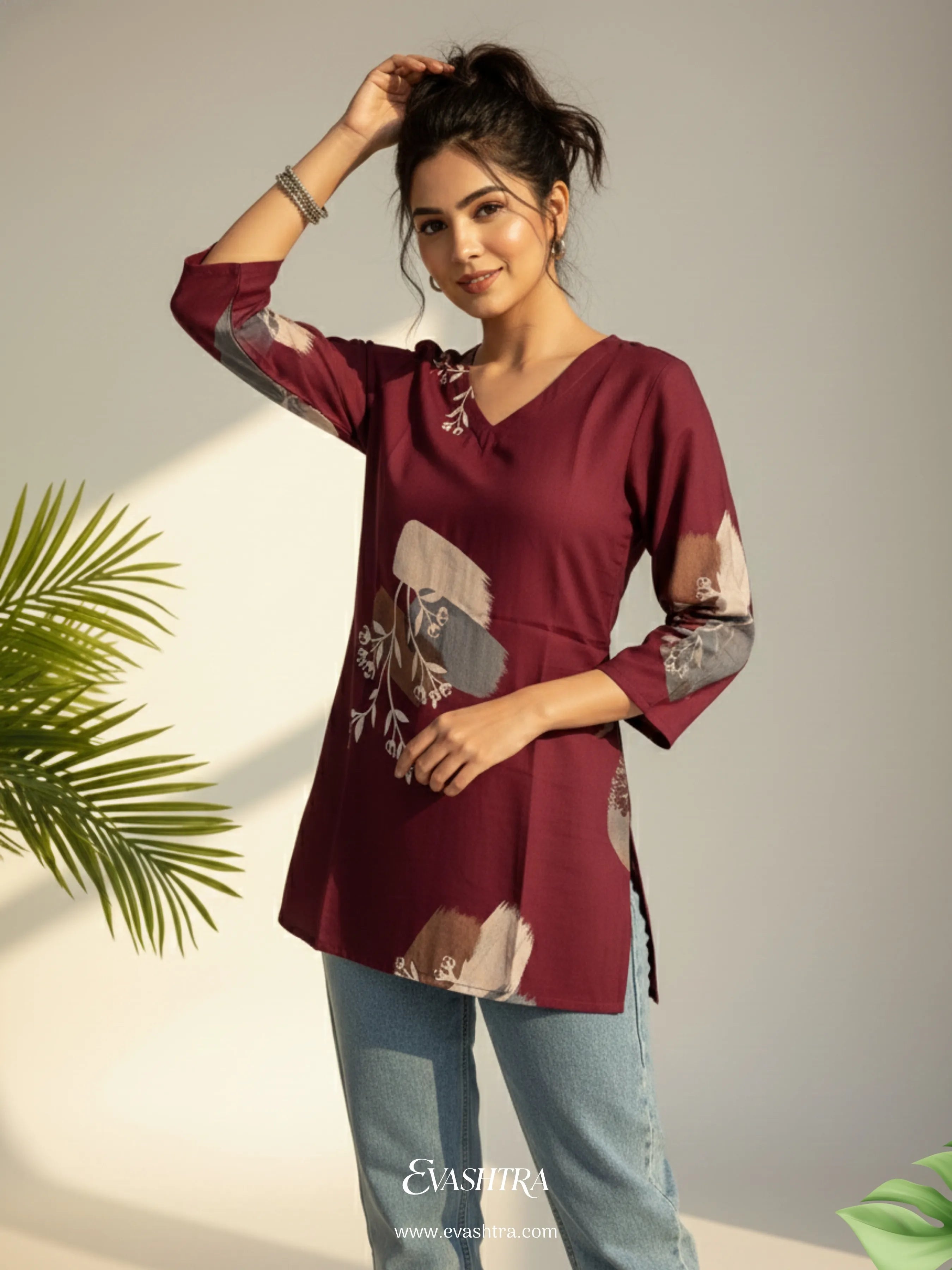 Woman wearing a maroon top with floral patterns and light blue jeans, standing against a neutral background.