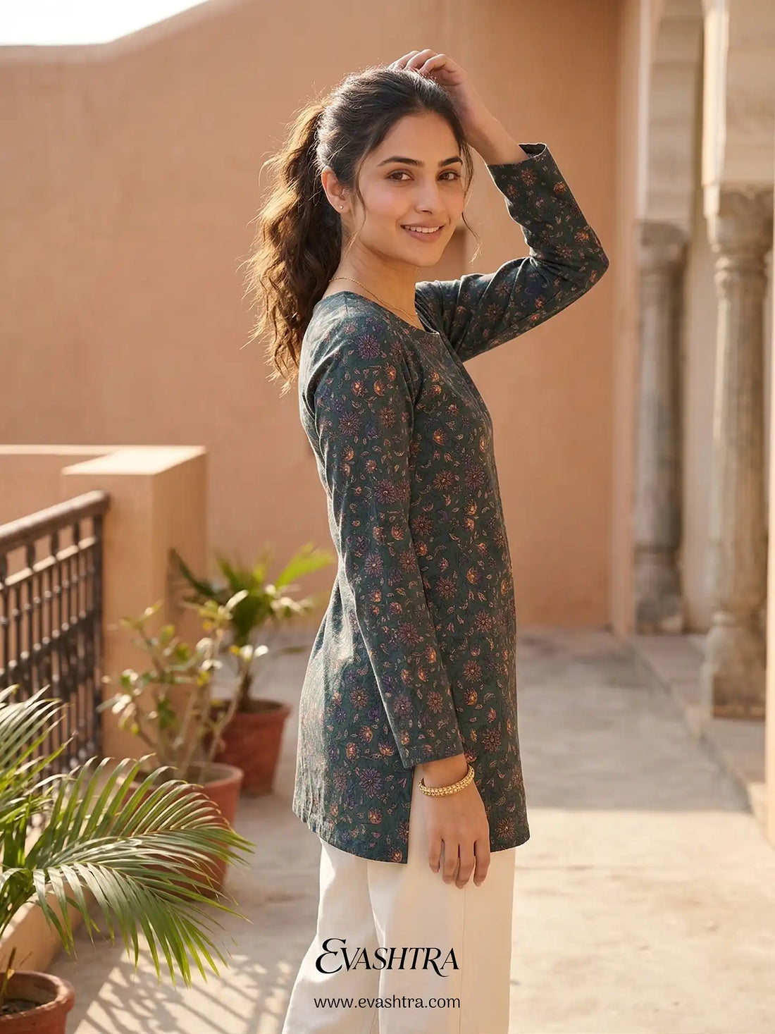 Woman wearing a dark green floral top and white pants standing outdoors with plants and a building in the background.