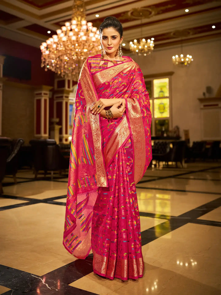 Woman in a pink saree with gold patterns standing in an elegant indoor setting.