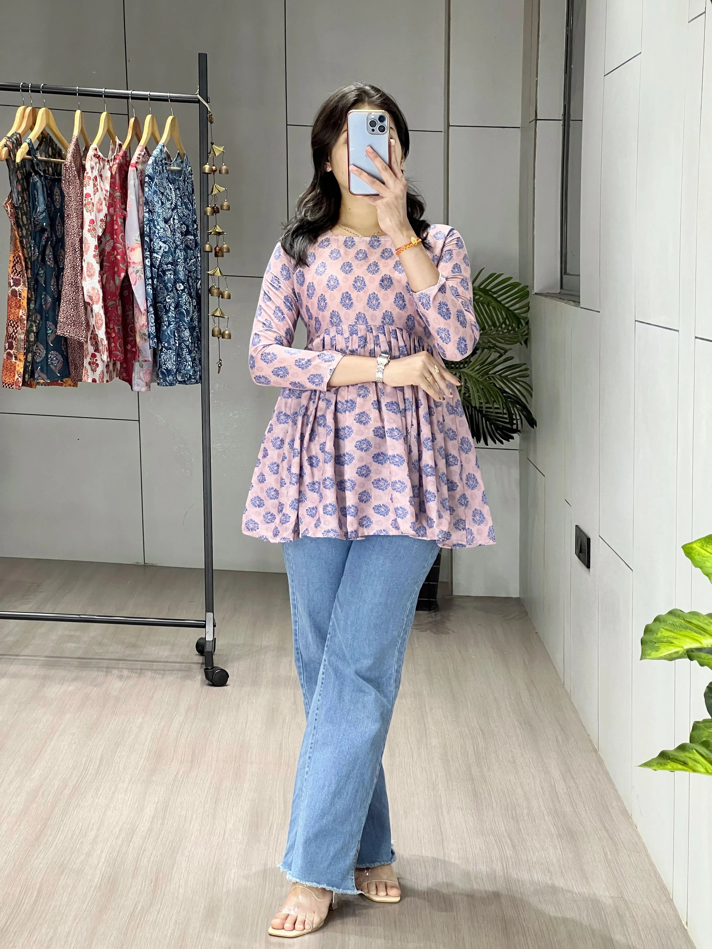 Woman taking a mirror selfie wearing a floral top and jeans in a room with a clothing rack.