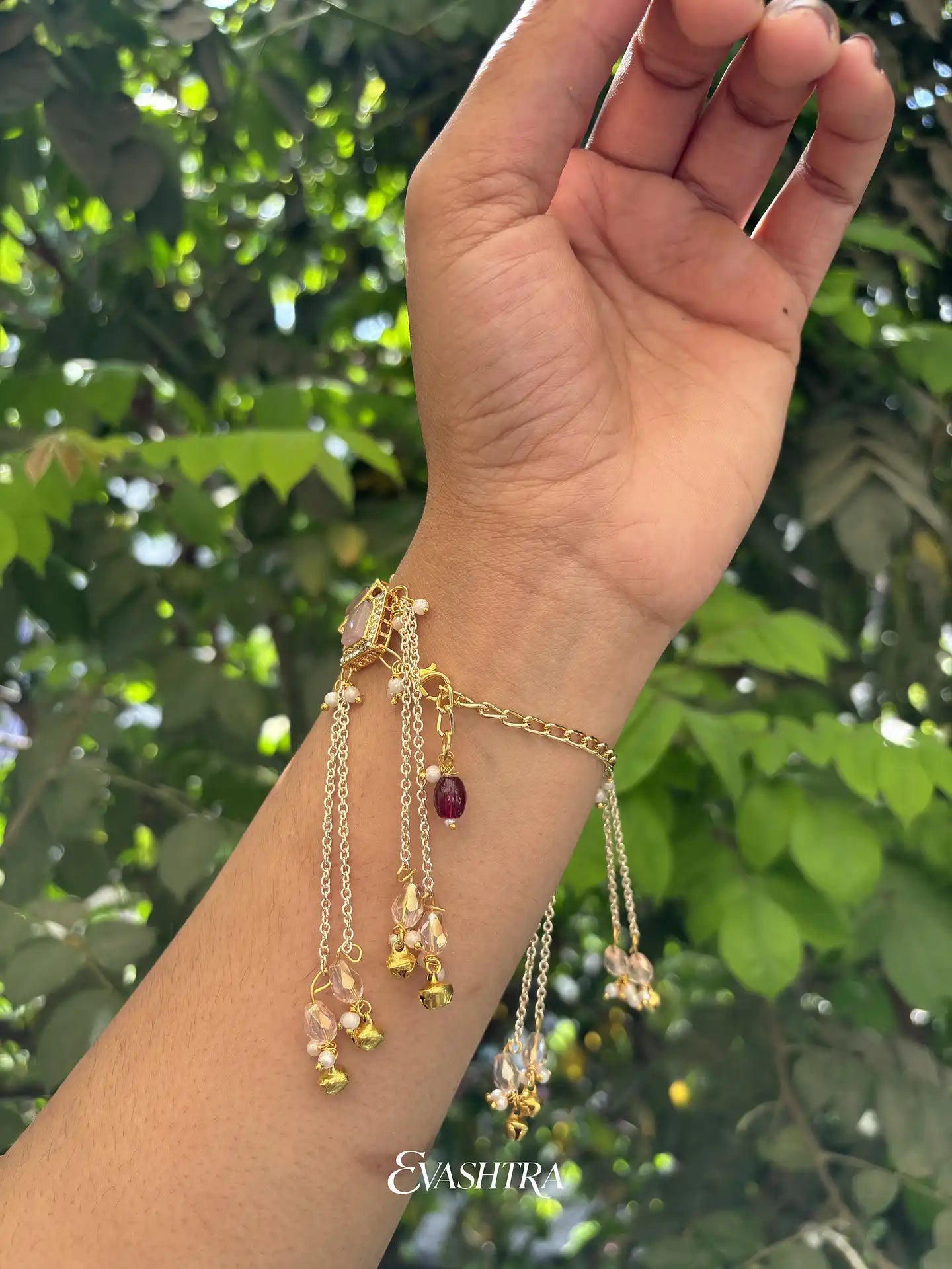 Hand wearing a gold bracelet with charms against a green leafy background