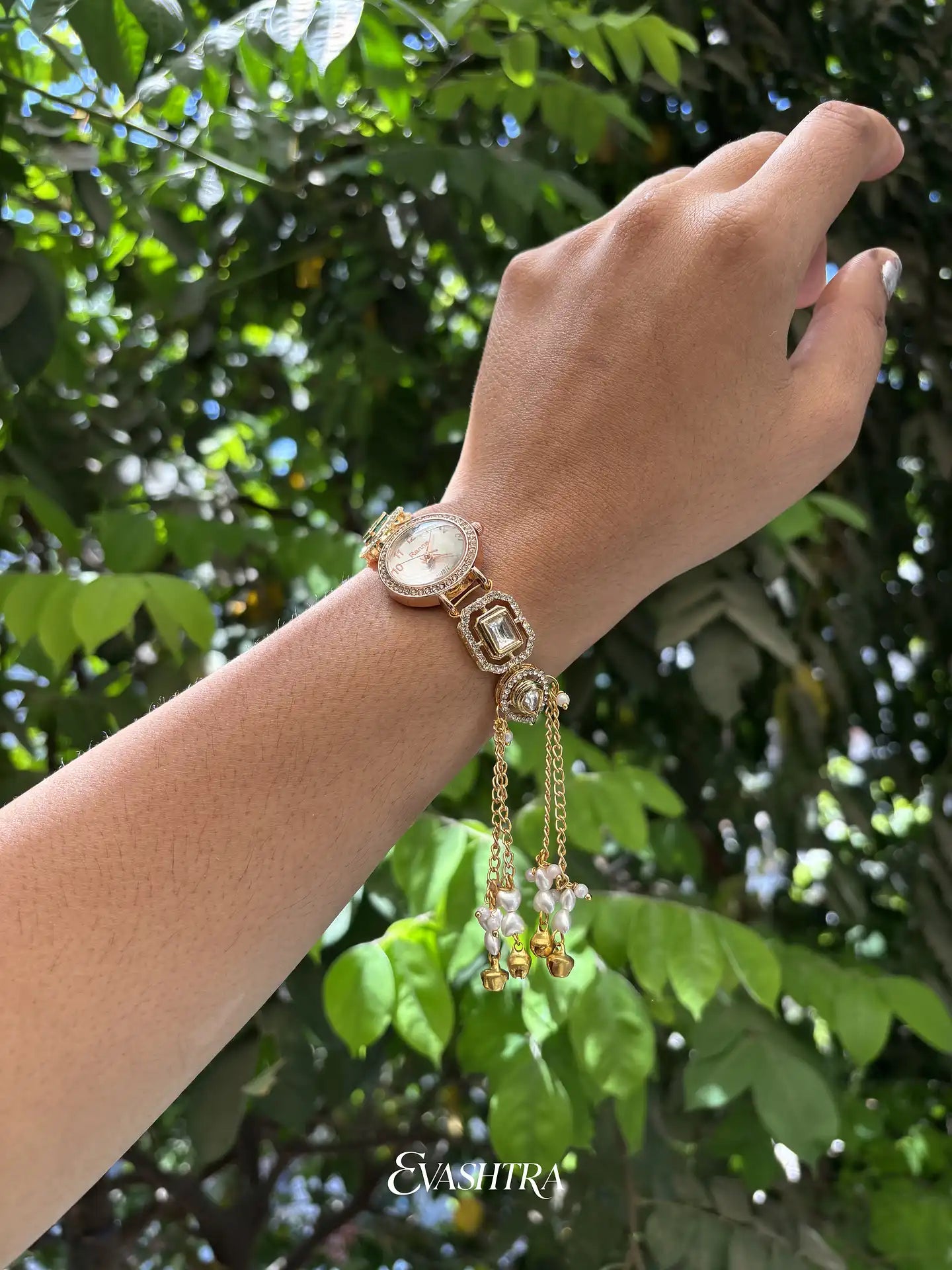 Hand wearing a gold bracelet with a watch face against a green leafy background