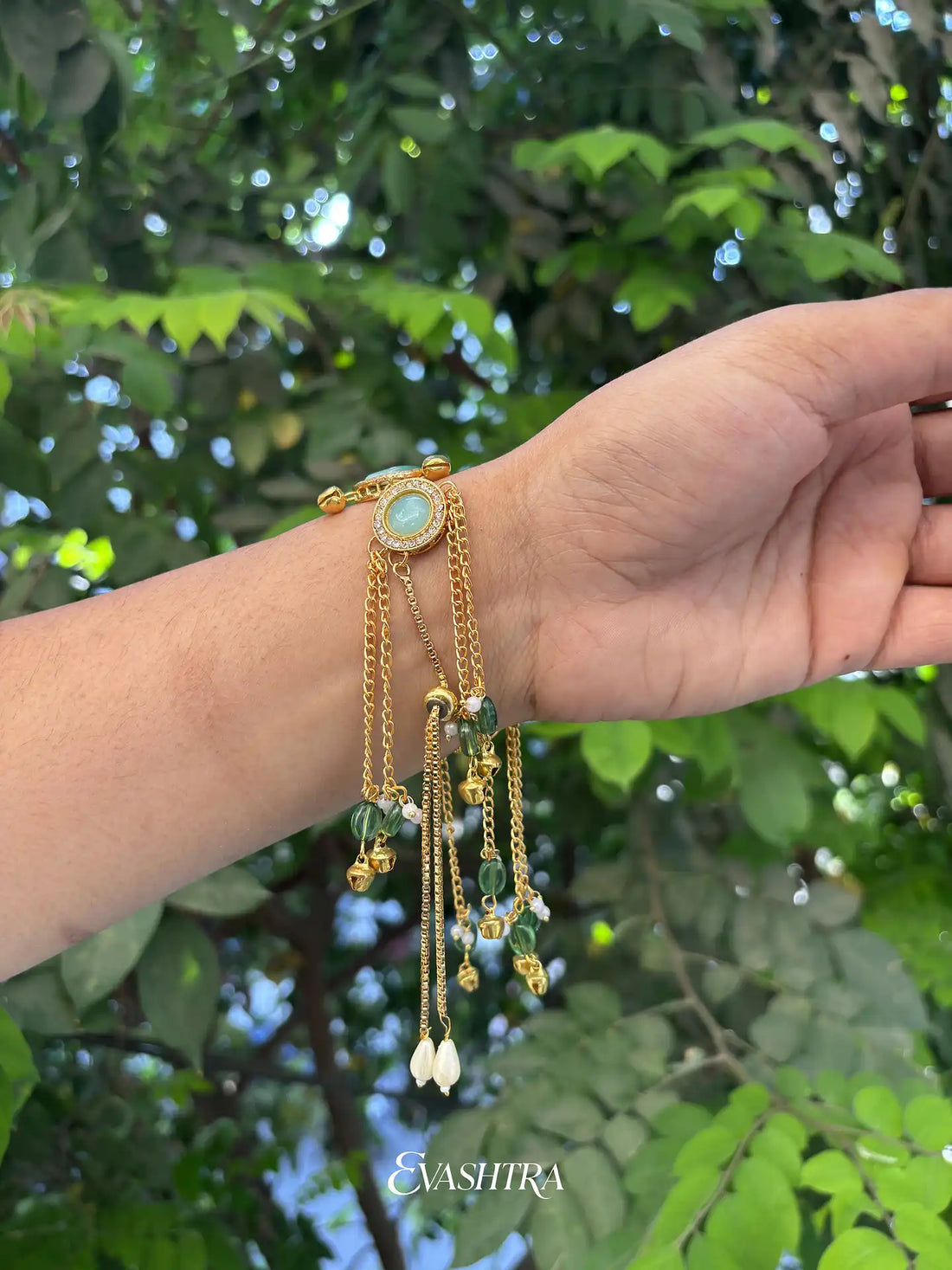 Hand wearing a gold bracelet with green stones against a green leafy background