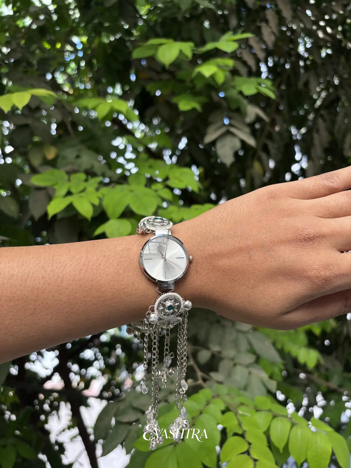Silver wristwatch with dangling charms on a wrist against a green leafy background