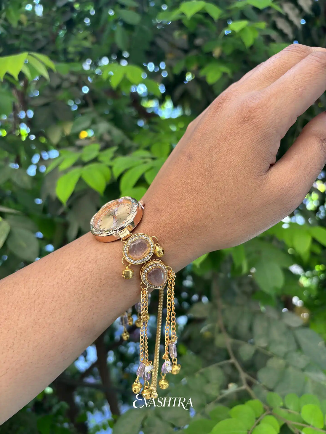 Hand wearing a gold bracelet with gemstones against a green leafy background