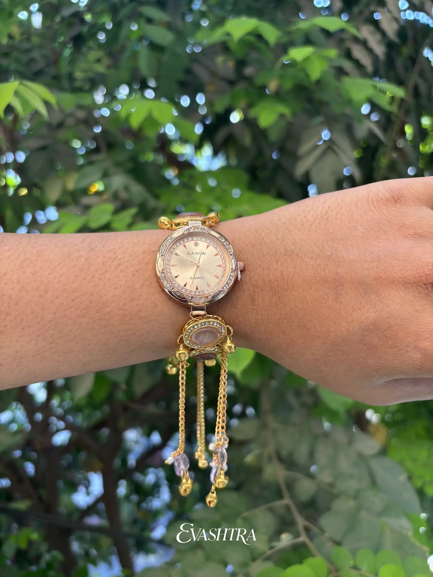 Gold wristwatch with decorative chain on a wrist against a green leafy background