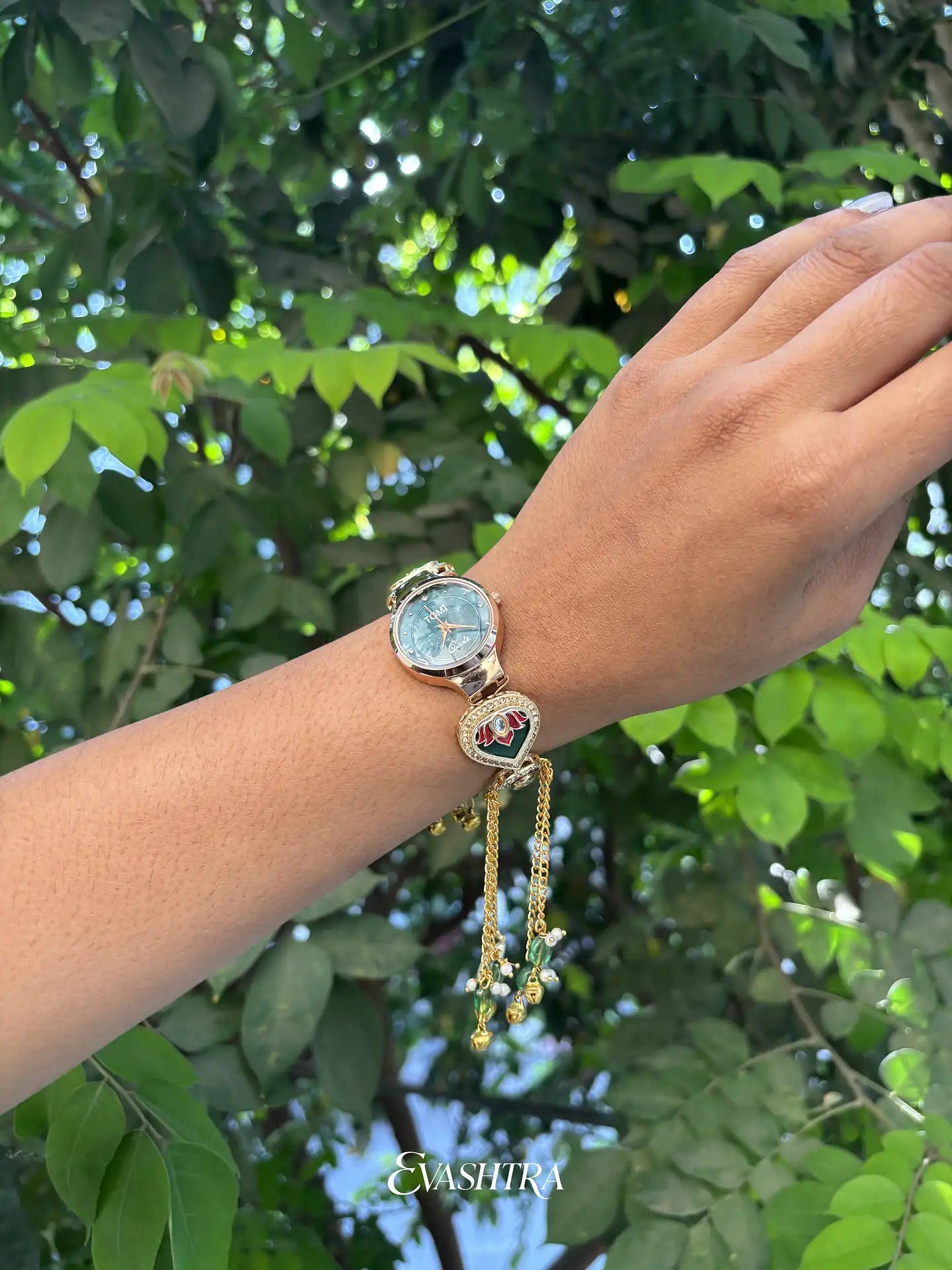 Hand wearing a gold bracelet with a watch face against a green leafy background