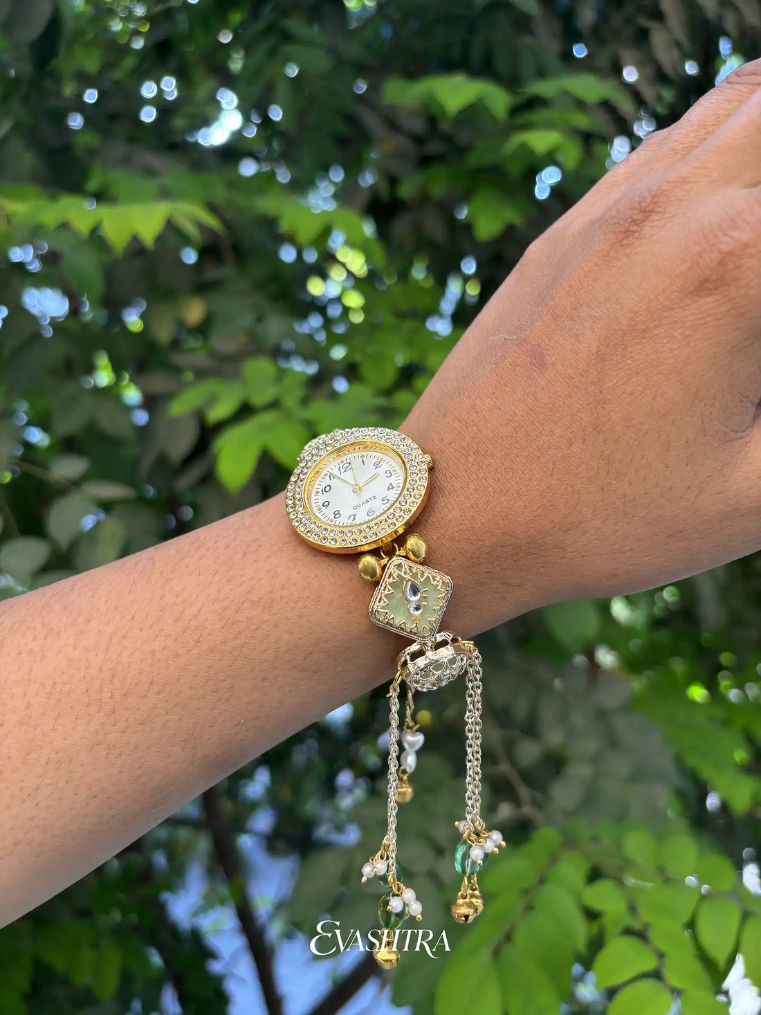 Gold wristwatch with decorative chain on a wrist against a green leafy background