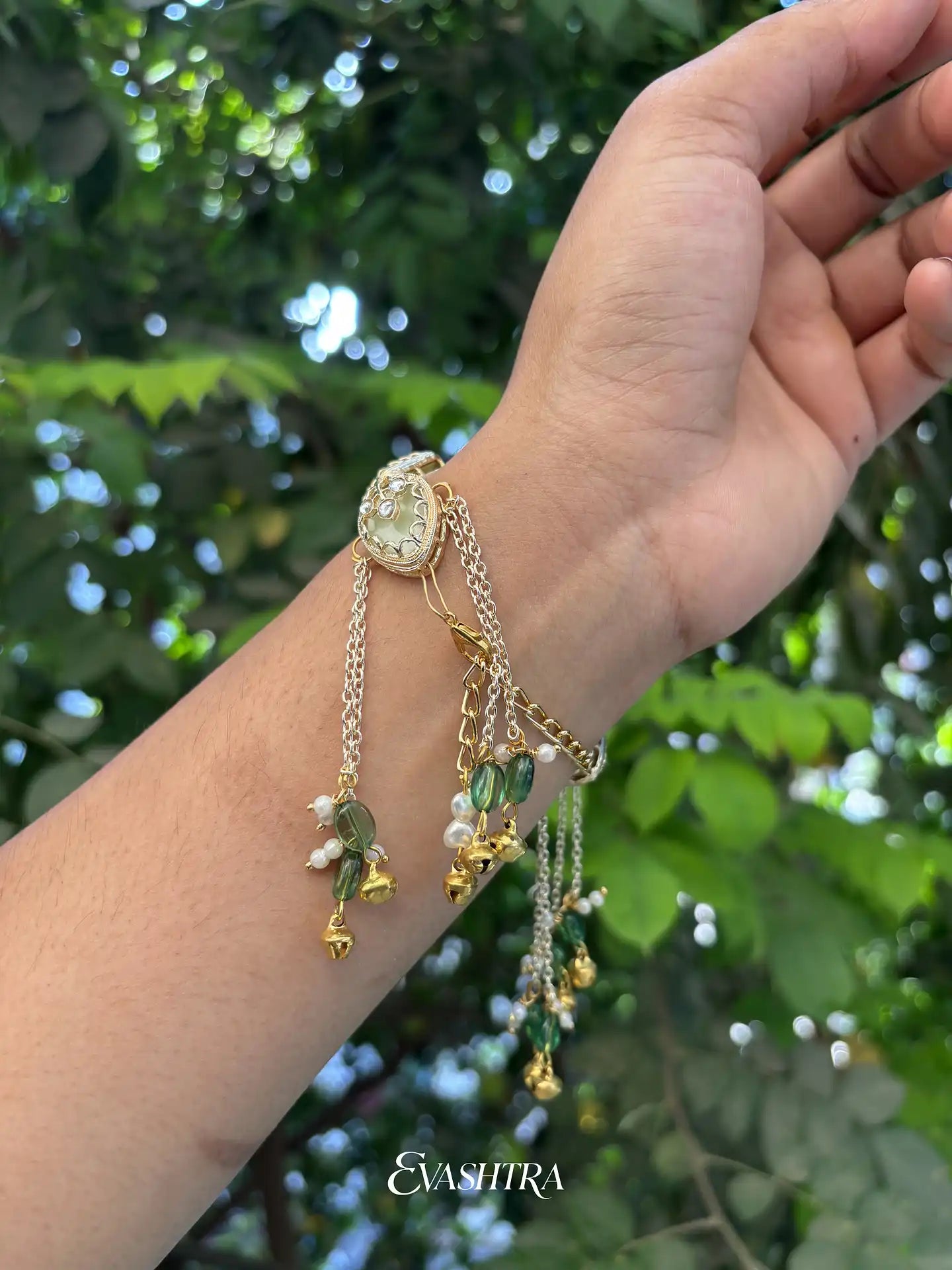 Hand wearing multiple gold bracelets with green and white beads against a blurred green leafy background