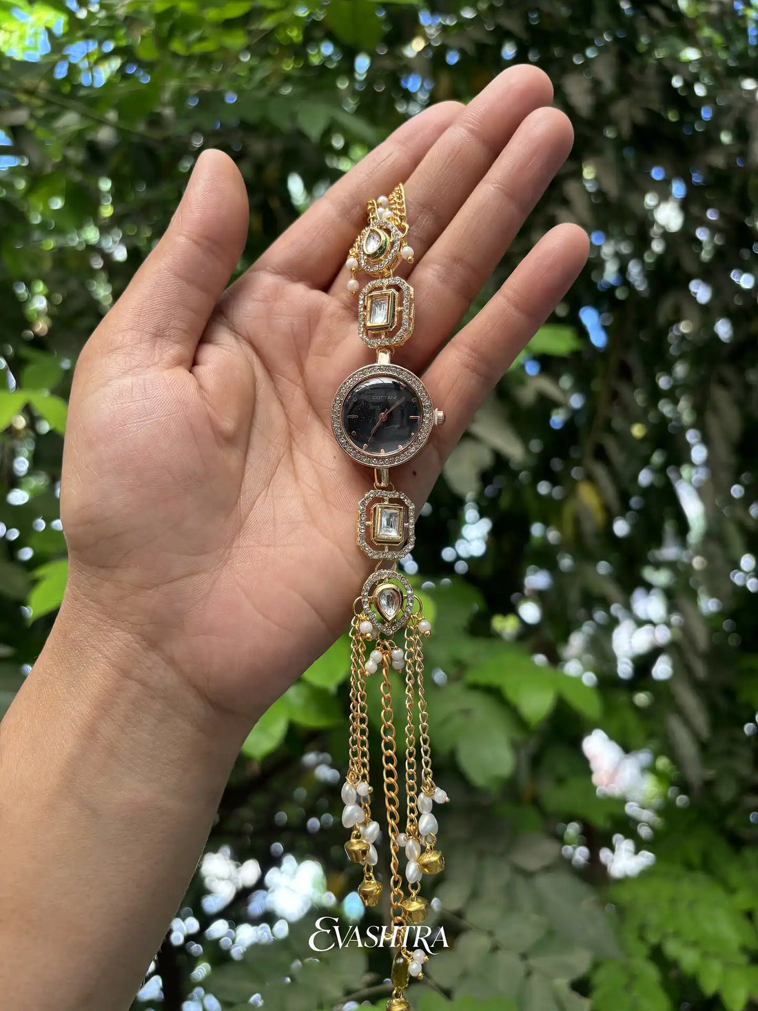 Hand holding a decorative gold and black jewelry piece with a blurred green foliage background