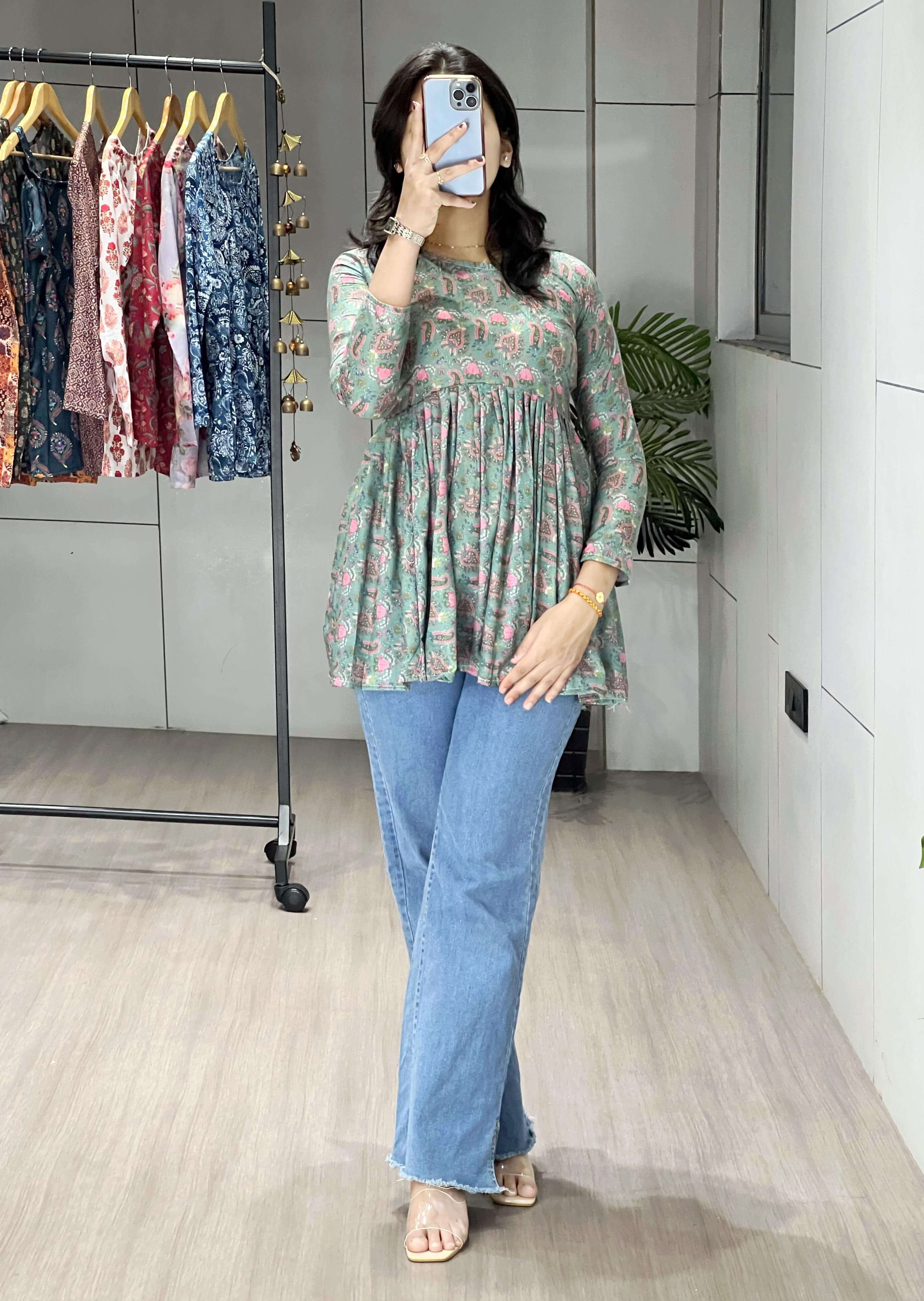 Woman taking a mirror selfie wearing a floral top and blue jeans in a room with a rack of clothes.